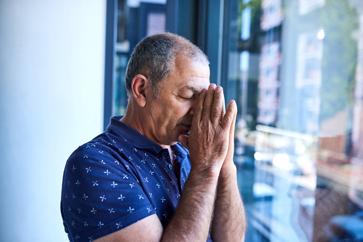 Close-up of a man praying with his hands clasped in front of his face looking out a building window.