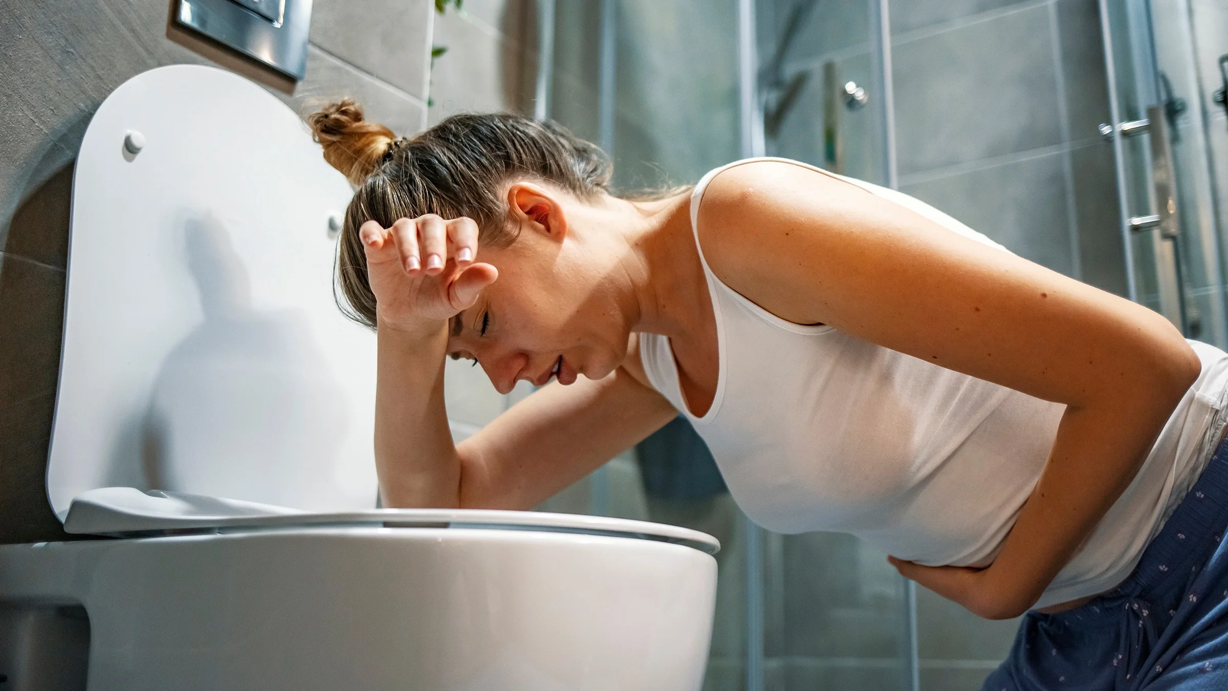 Young woman hunched over the toilet about to throw up. She is clutching her stomach with one hand and using the back of her other arm to rest her head on.