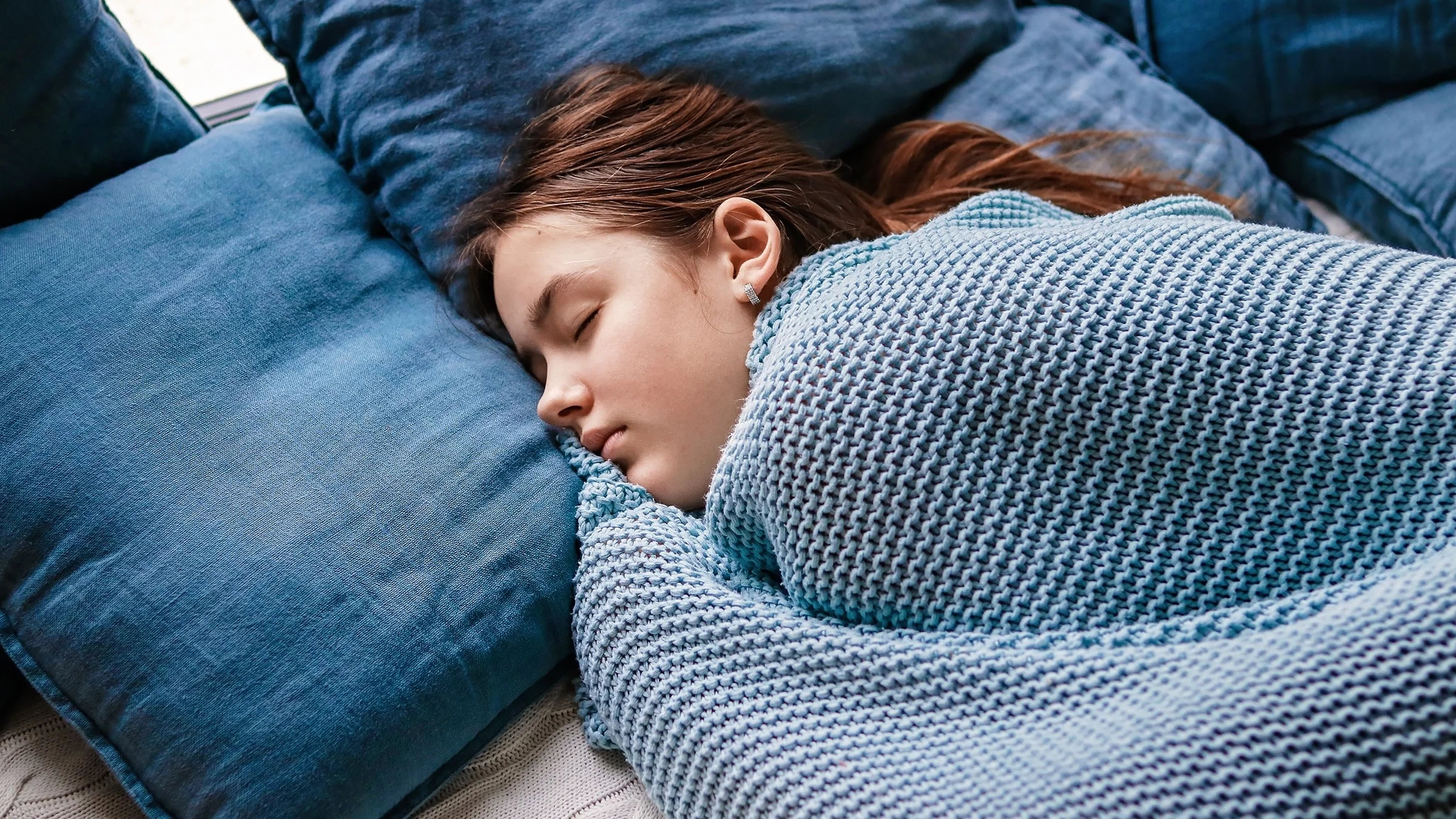 Teenage girl sleeping in bed. She's bundled up with a crocheted blue blanket and blue pillows.
