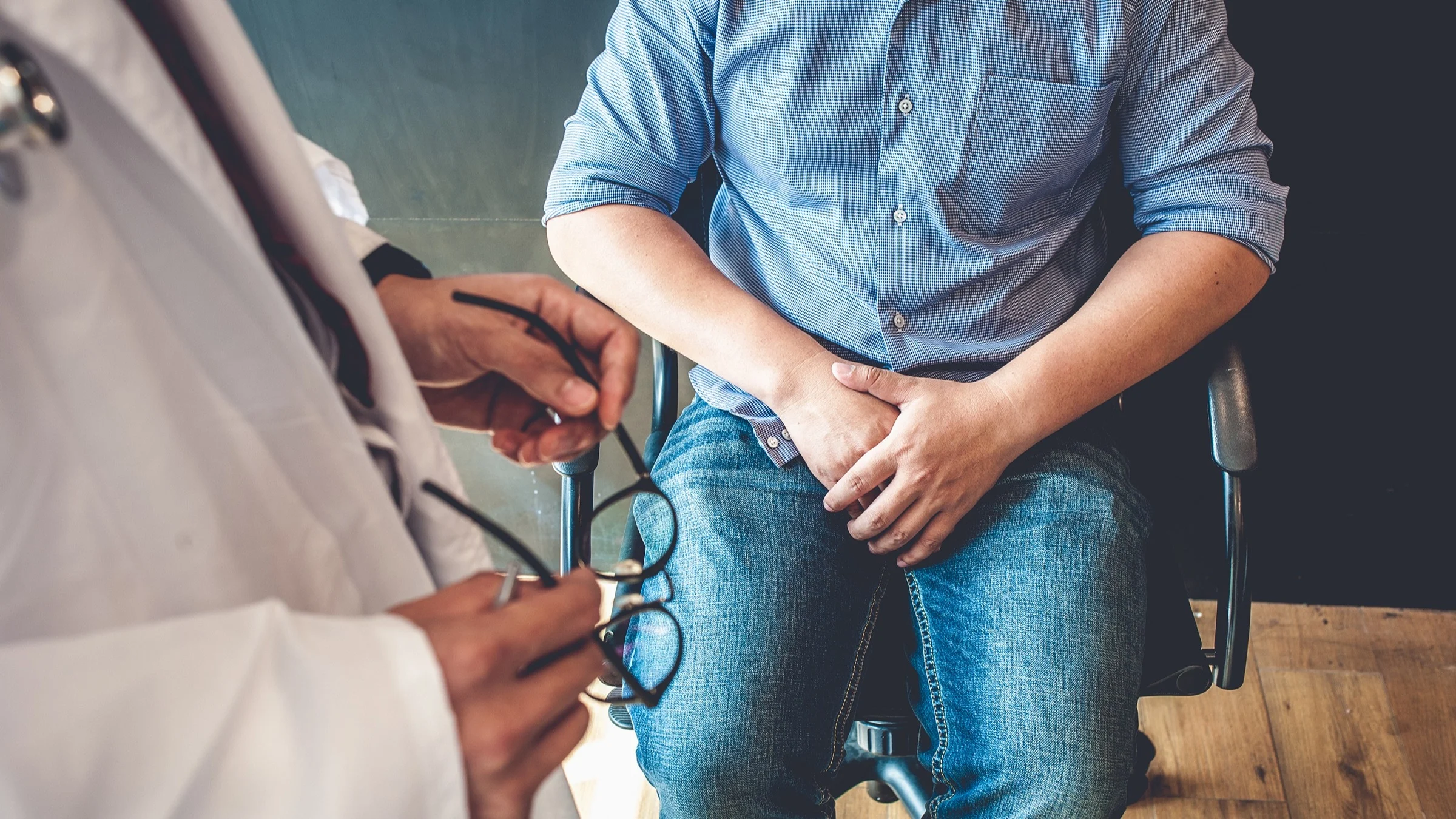 Cropped shot of a man sitting in a swivel chair covering his lap with his hands. The doctor is standing up next to him holding their glasses in their hands.