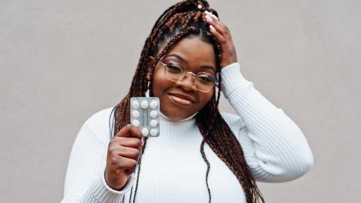 Portrait of a young Black woman holding up a blister pack of pills on a textured gray background.