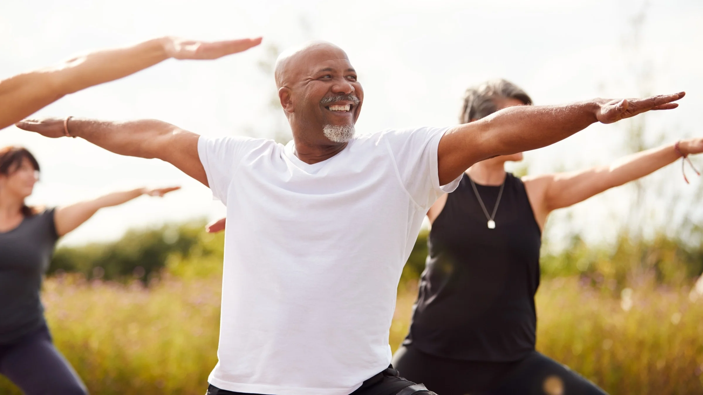 Older man doing yoga in a field with a group of women. They are in warrior pose.