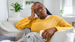 Woman with eyes closed and hand on forehead sitting on the couch with a blanket.
stefanamer/iStock via Getty Images