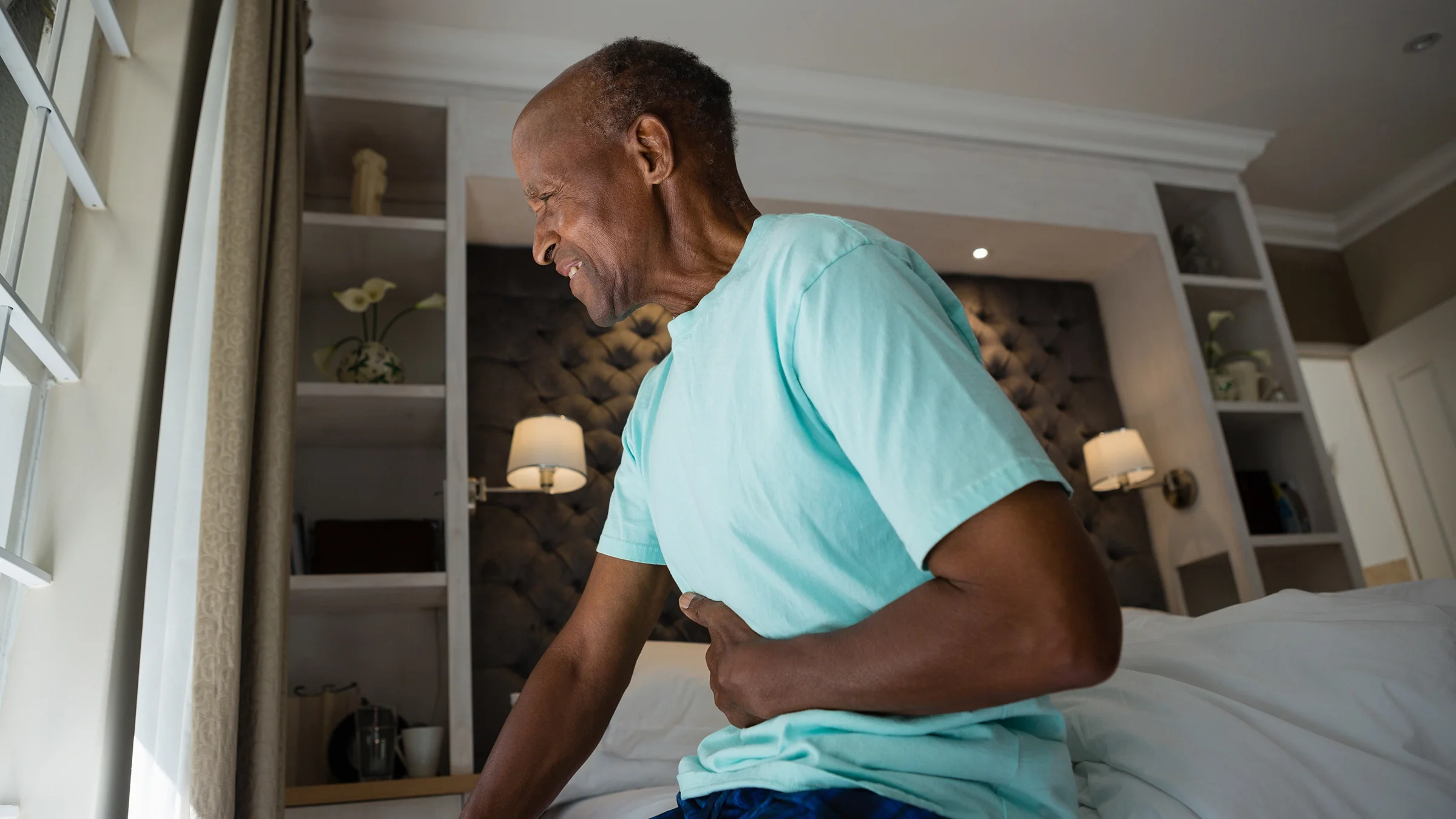 An older man suffering from stomach ache while sitting on a bed at home.