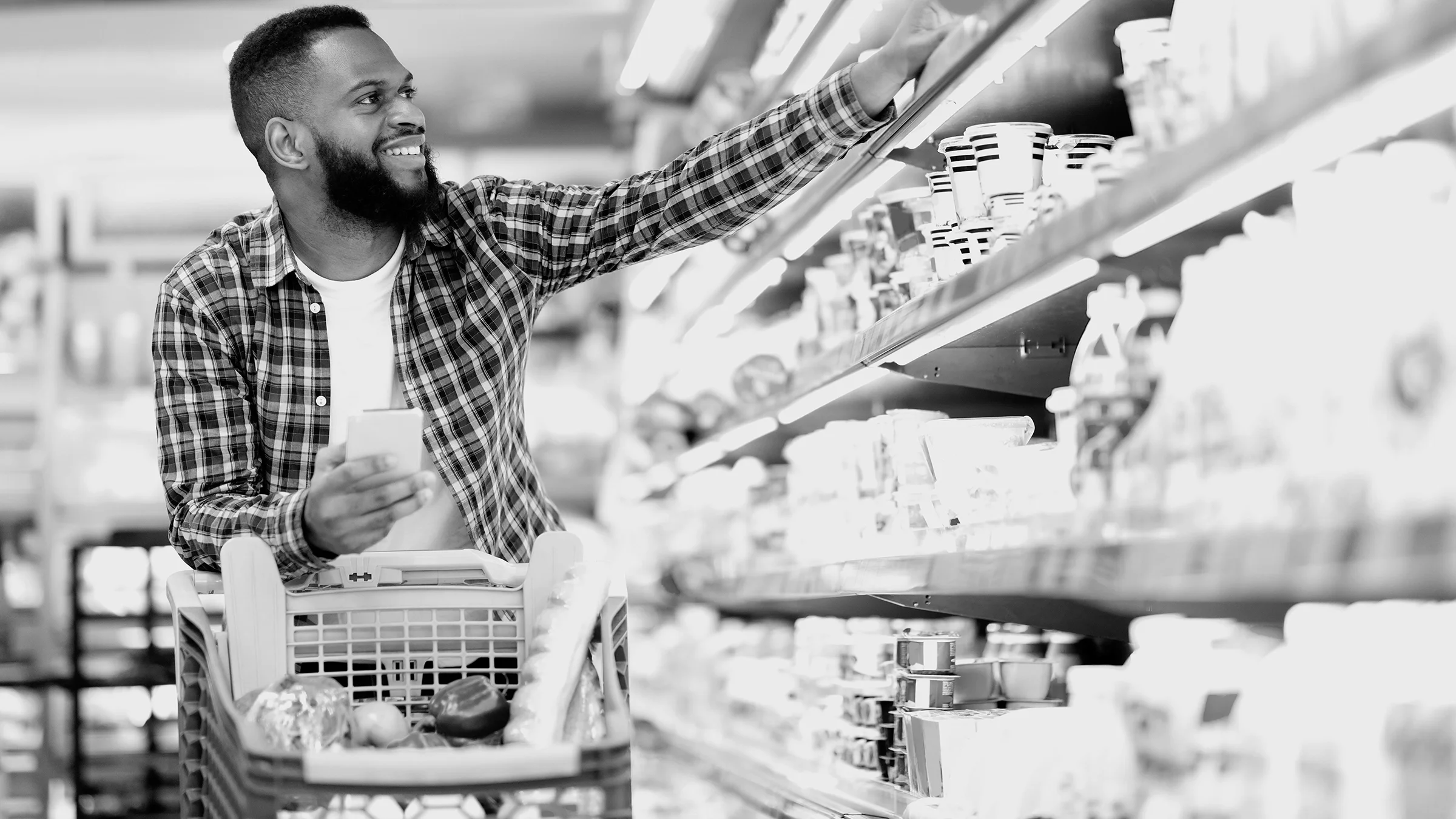 Black and white photo of a man grocery shopping in the yogurt aisle. He is reaching up to the top shelf.