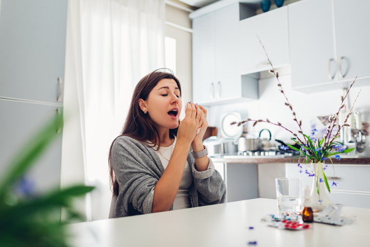 Woman sneezing at her kitchen table with flowers in the middle.