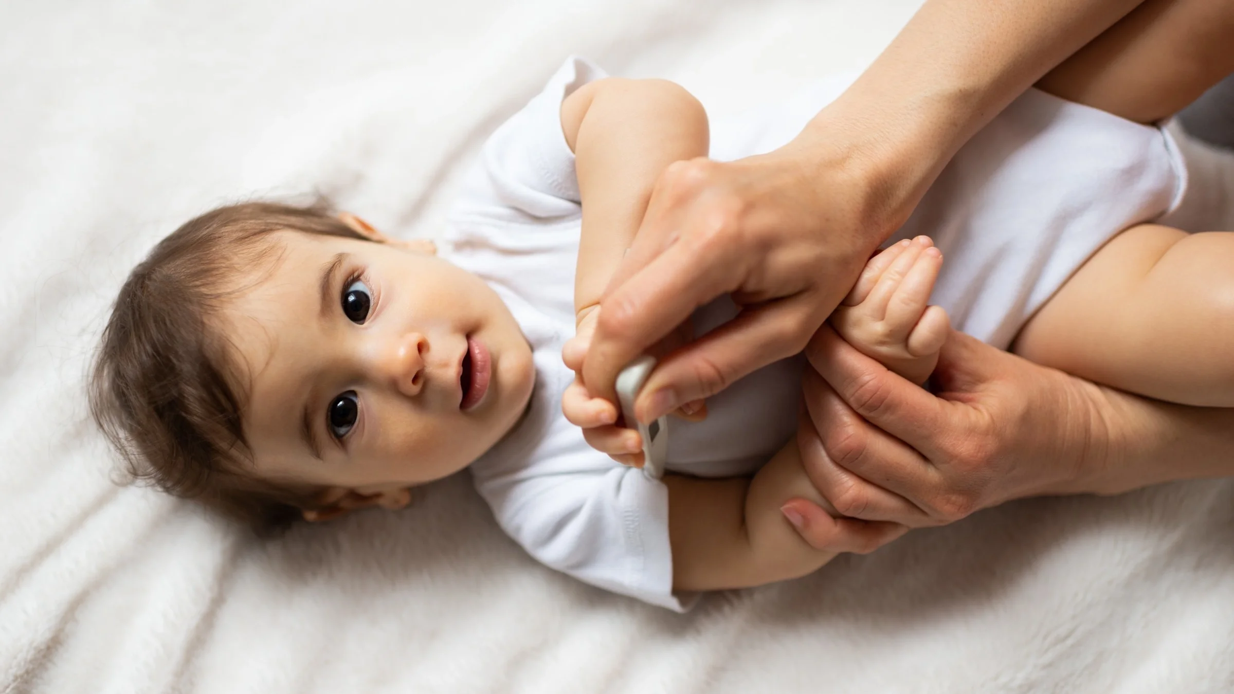 Close-up portrait of a baby in a white onesie while the mother checks temperature with a digital thermometer.