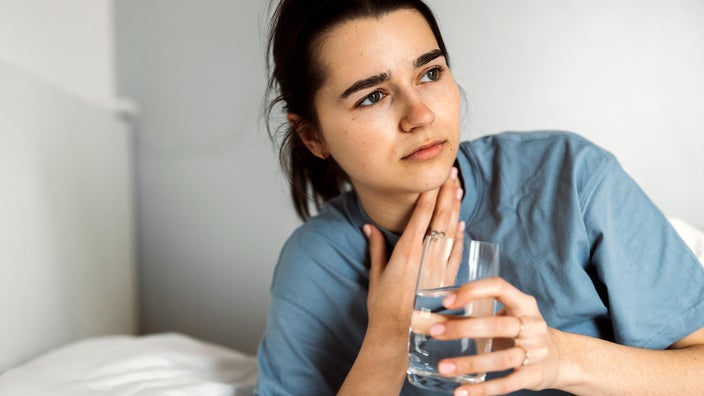 A woman with a sore throat rests in bed while holding a glass of water. Tonsil stones can cause pain in the throat.