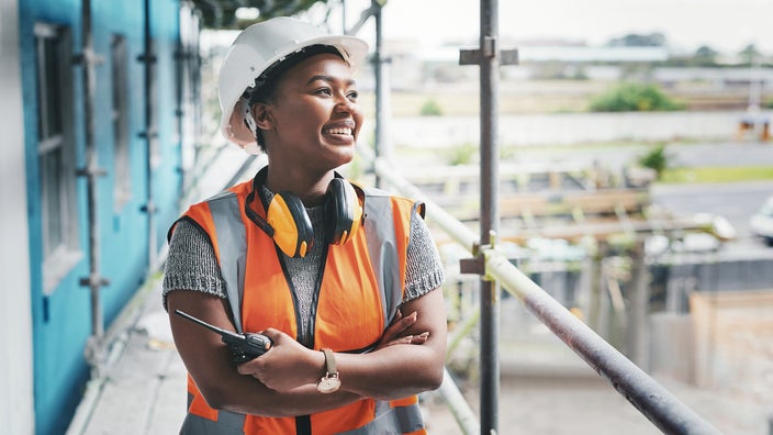 Young woman working at a construction site. She is on the scaffolding and wearing a hard hat and orange safety vest.