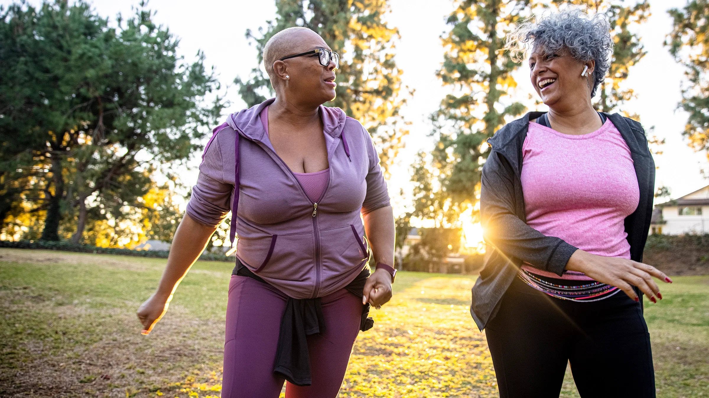 Two women on a walk through the park with workout clothes on. They are smiling and laughing together like best friends.