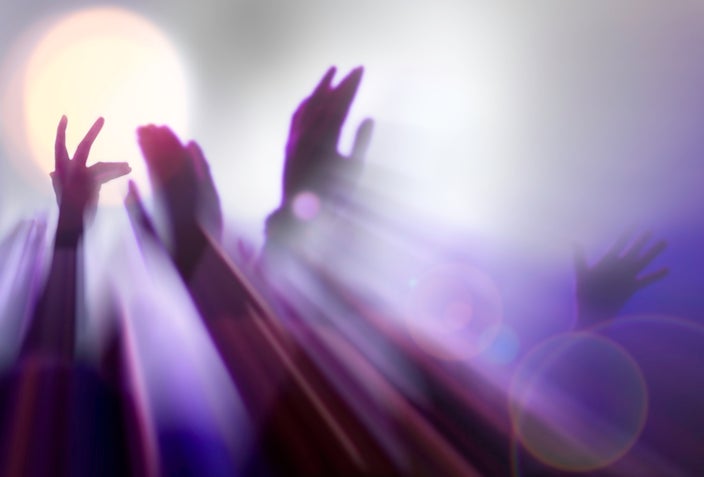 Hands in the air at a concert or event. There is a big bright lens flare and the frame is tinted purple.