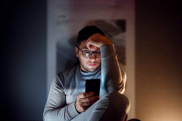 Portrait of a man sitting in the dark looking at social media on his phone. He looks depressed and frustrated.