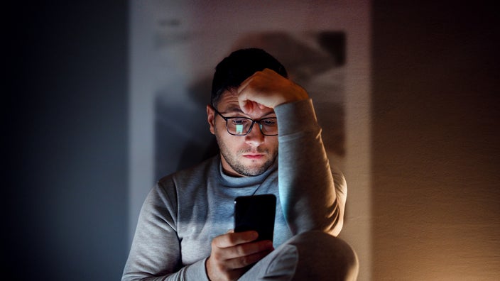 Portrait of a man sitting in the dark looking at social media on his phone. He looks depressed and frustrated.