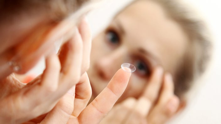 A person inserting a contact lens in their eye in the mirror. The finger with the lens is in focus.