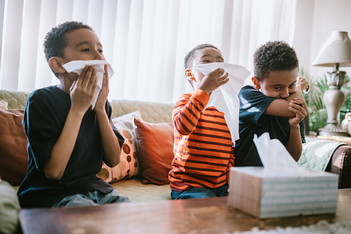 Three young boys sitting on the couch blowing their noses and coughing with tissue box on the coffee table.