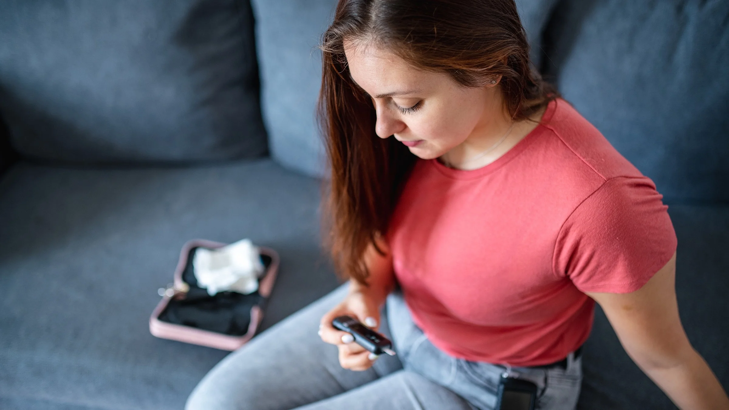 Mujer joven con cabello largo sentada en el sofá comprobando su nivel de azúcar en la sangre. También lleva un medidor de glucosa en la cadera.