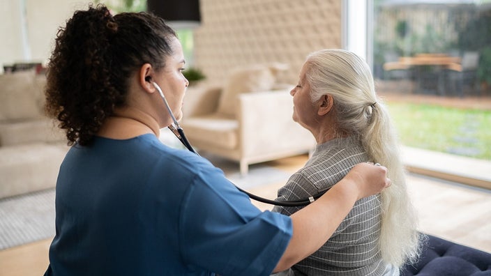 A nurse listening to a patient's heart beat with the stethoscope on the patient's back during a home check-in.