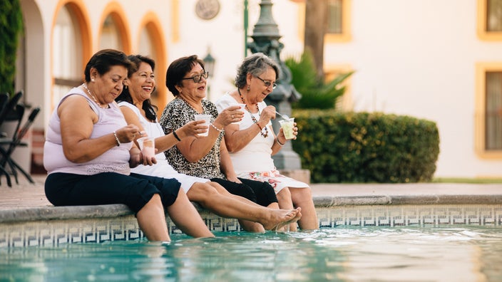A group of elderly woman dipping their feet in the pool while drinking margaritas.