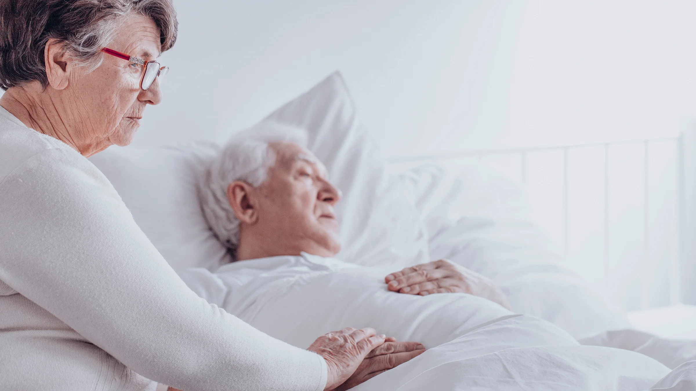 An older adult is sitting at the bedside next to her husband who is sick.