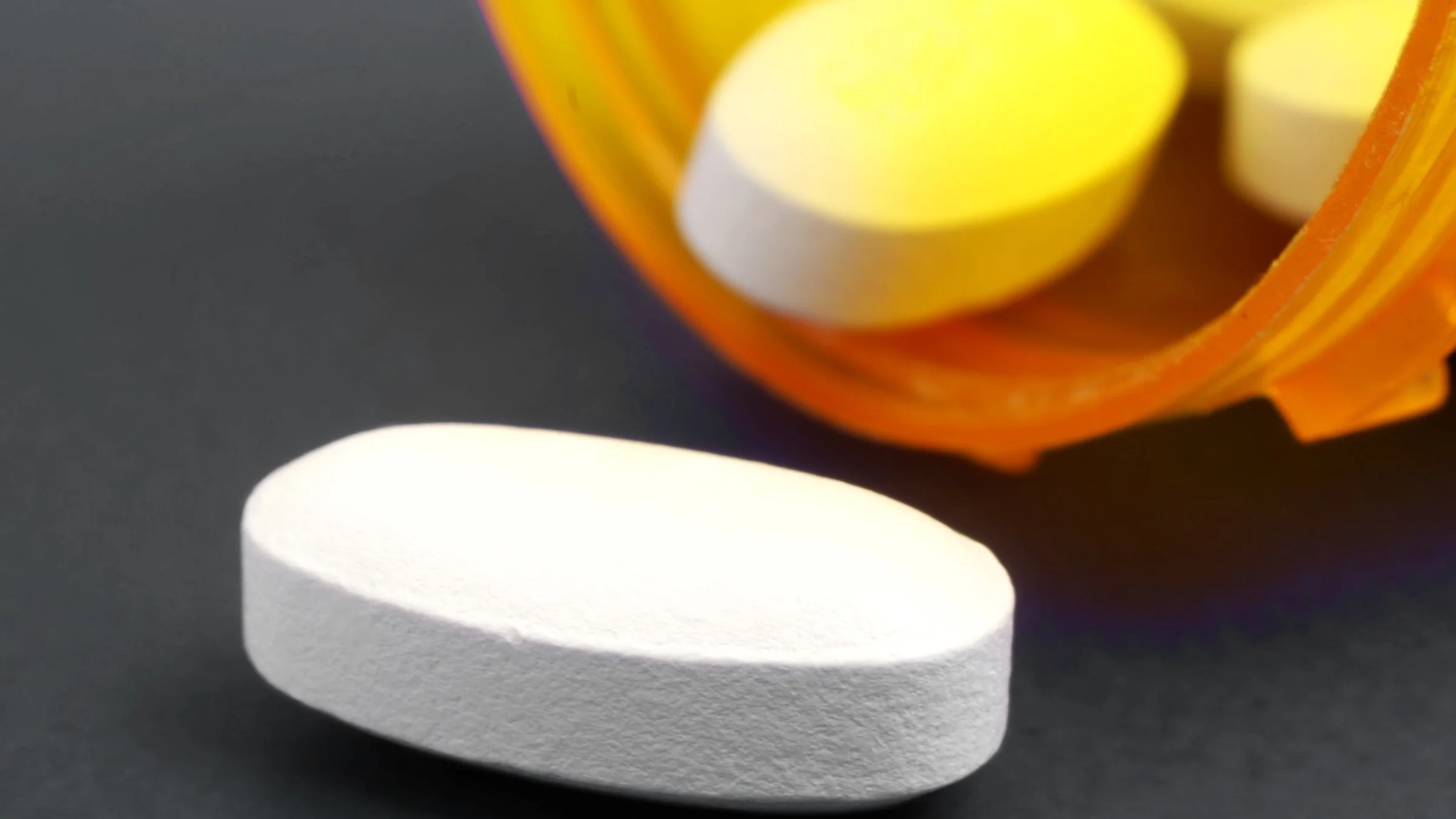 Oblong white pill spilling out of an orange prescription bottle on a black and gray background.