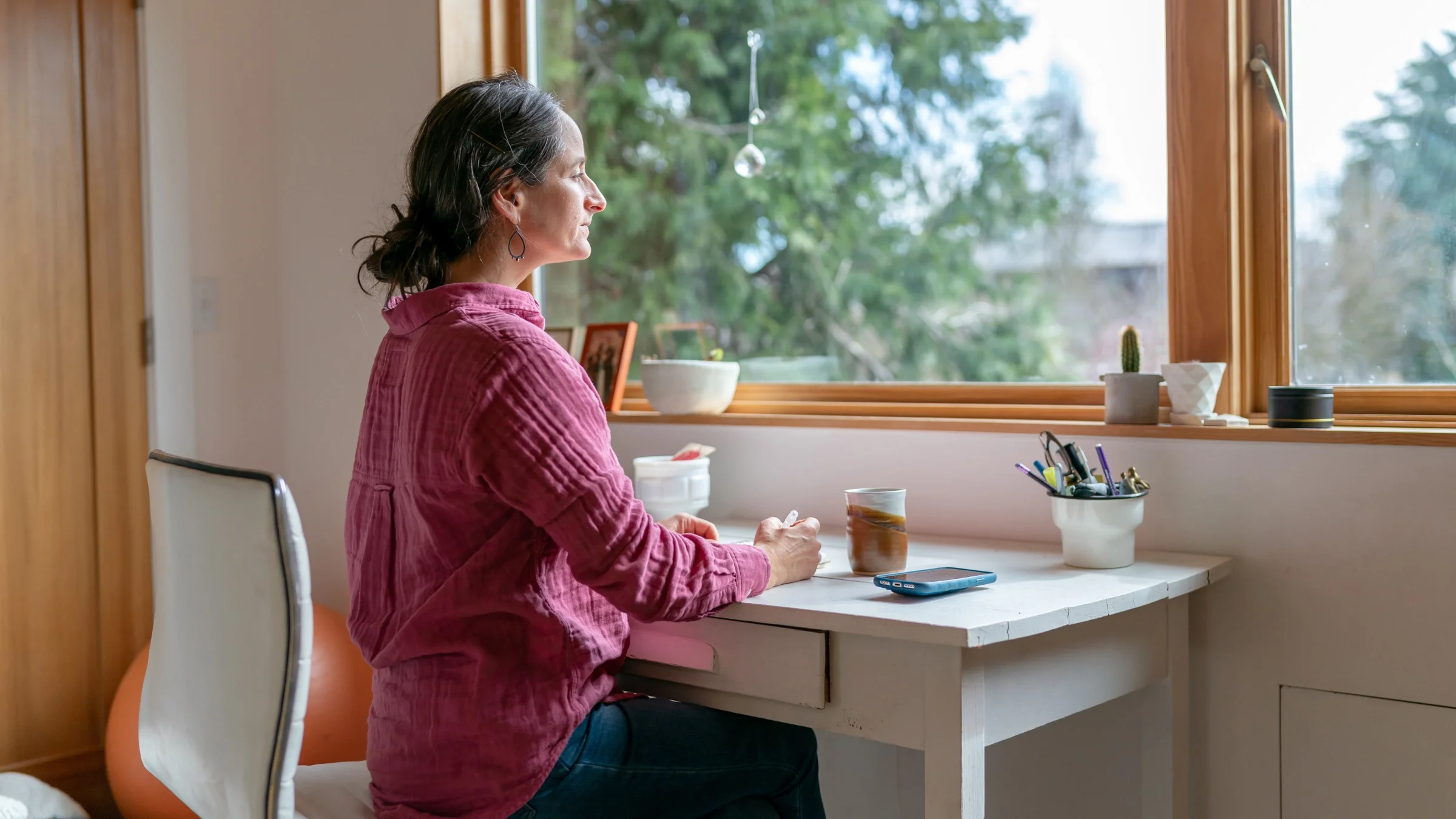Woman sits looking out the window with a contemplative expression while writing in a journal or diary.