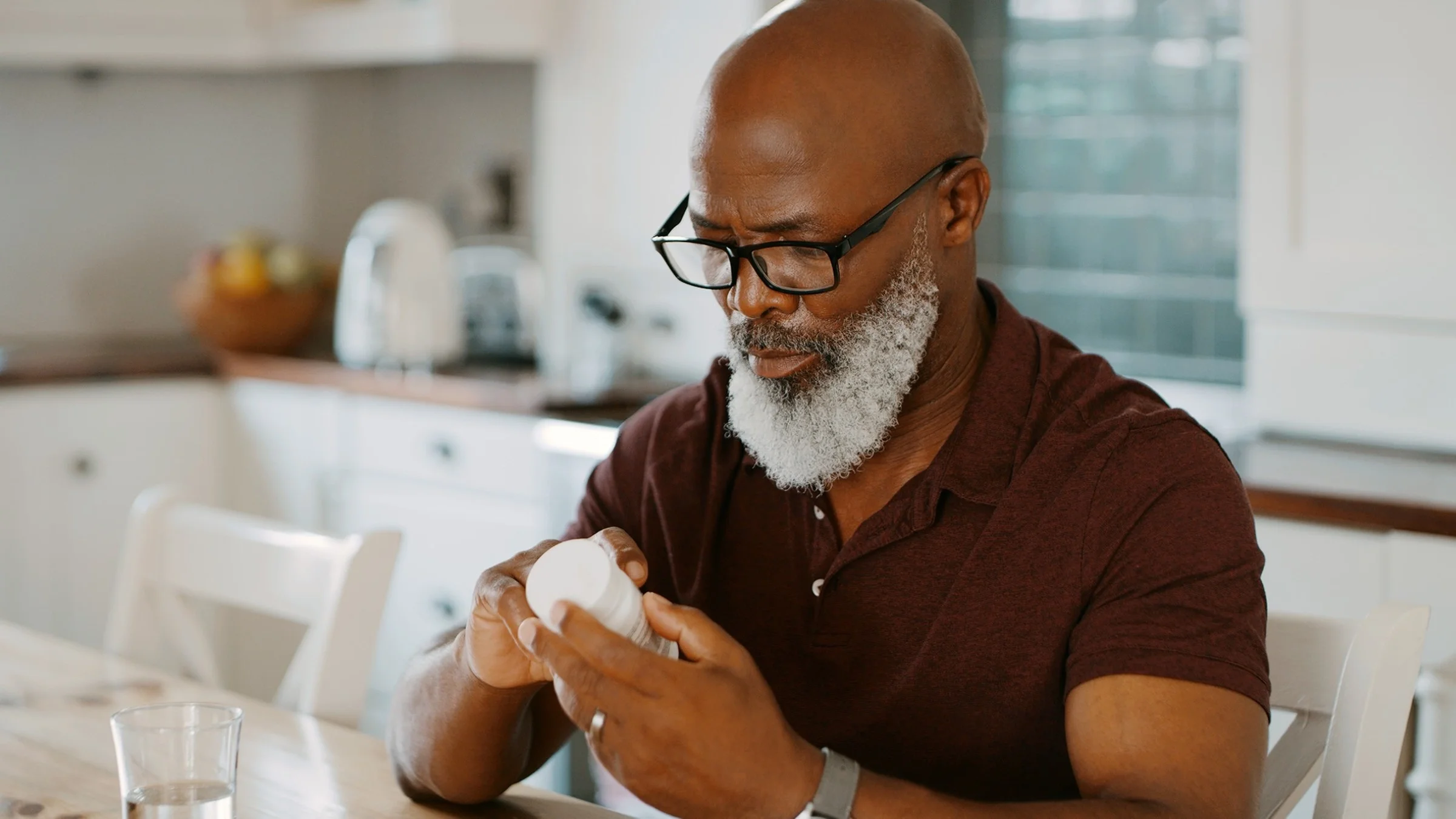 Senior man who is bald with a long gray beard reading the label of a prescription bottle. He is in his kitchen at the dining table.