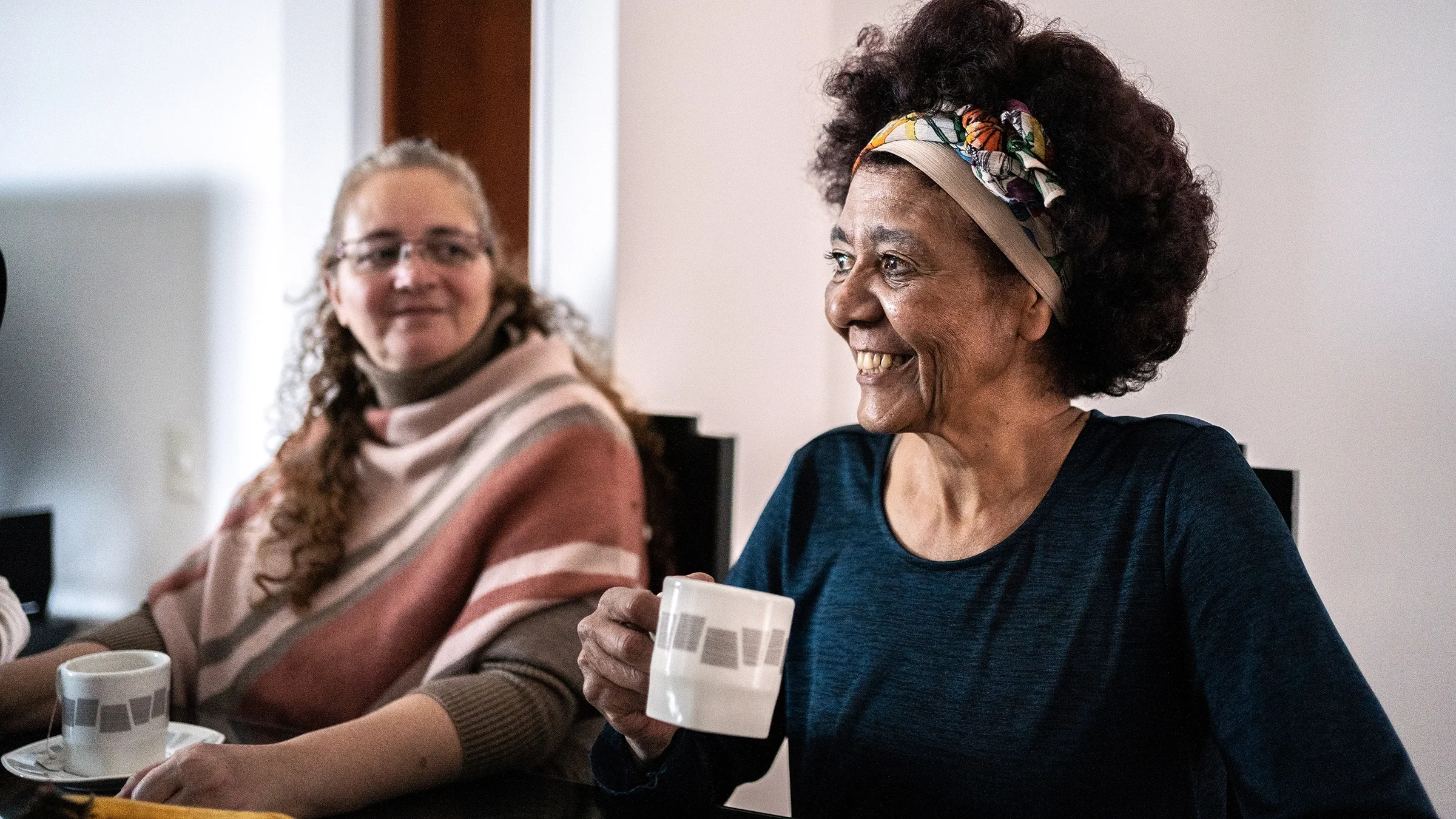A senior woman talks with her friends at home while holding a mug. 