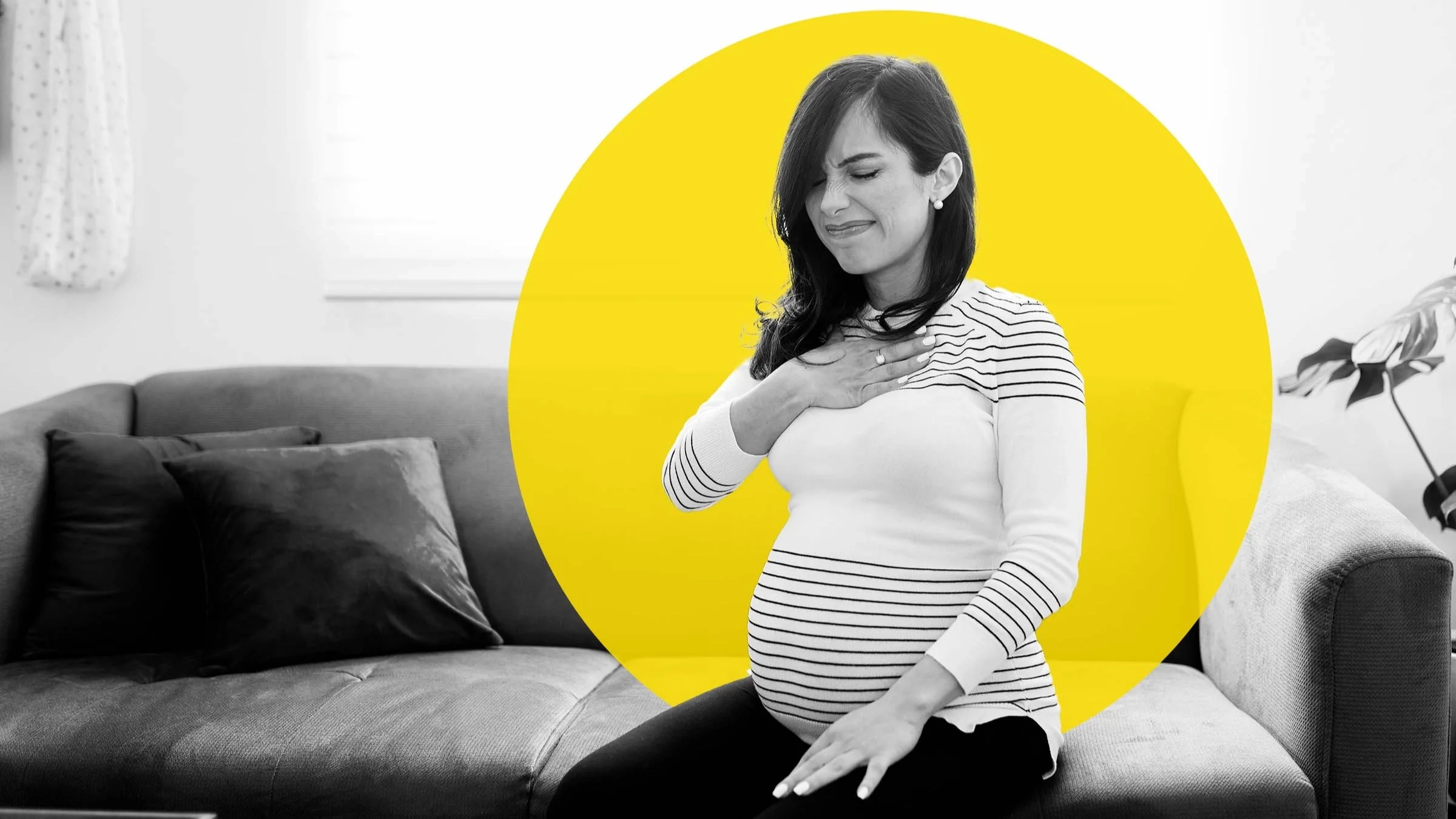 Black and white photo of a pregnant woman sitting on the couch with heartburn. She has her hand over her chest and is wincing in pain. There is an added yellow graphic circle behind her as well.