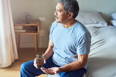 Older man taking medication with a glass of water, sitting on his bed looking out the window.
PeopleImages/E+ via Getty Images