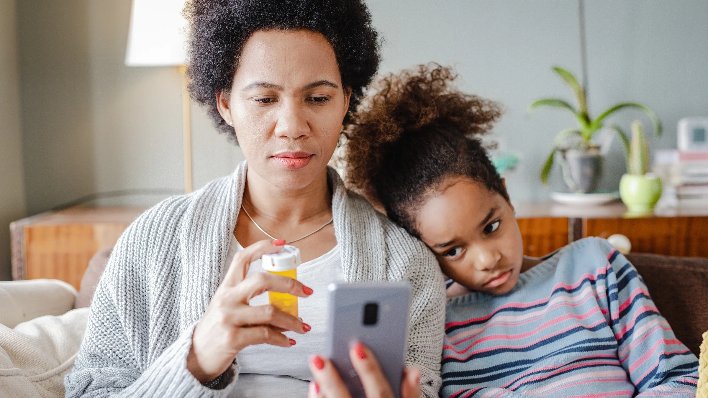 Mother and young daughter sitting on the couch using a mobile phone for a telehealth visit. The mother is holding a prescription pill bottle while the daughter rests her head on her mother's shoulder.