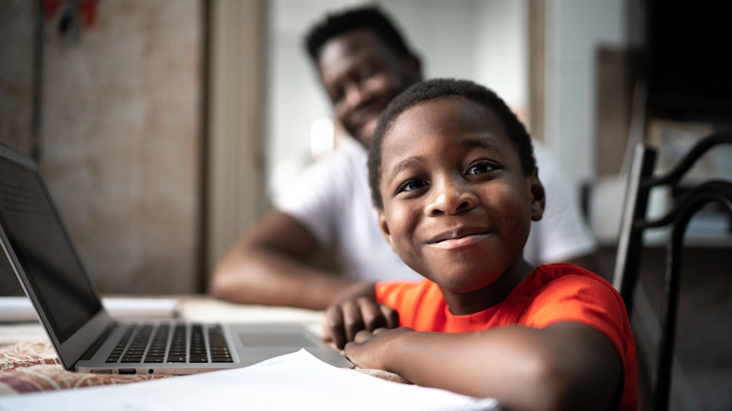 Portrait of a young boy smiling at the camera while he sits at a laptop doing homework. His dad is in the background and out of focus.