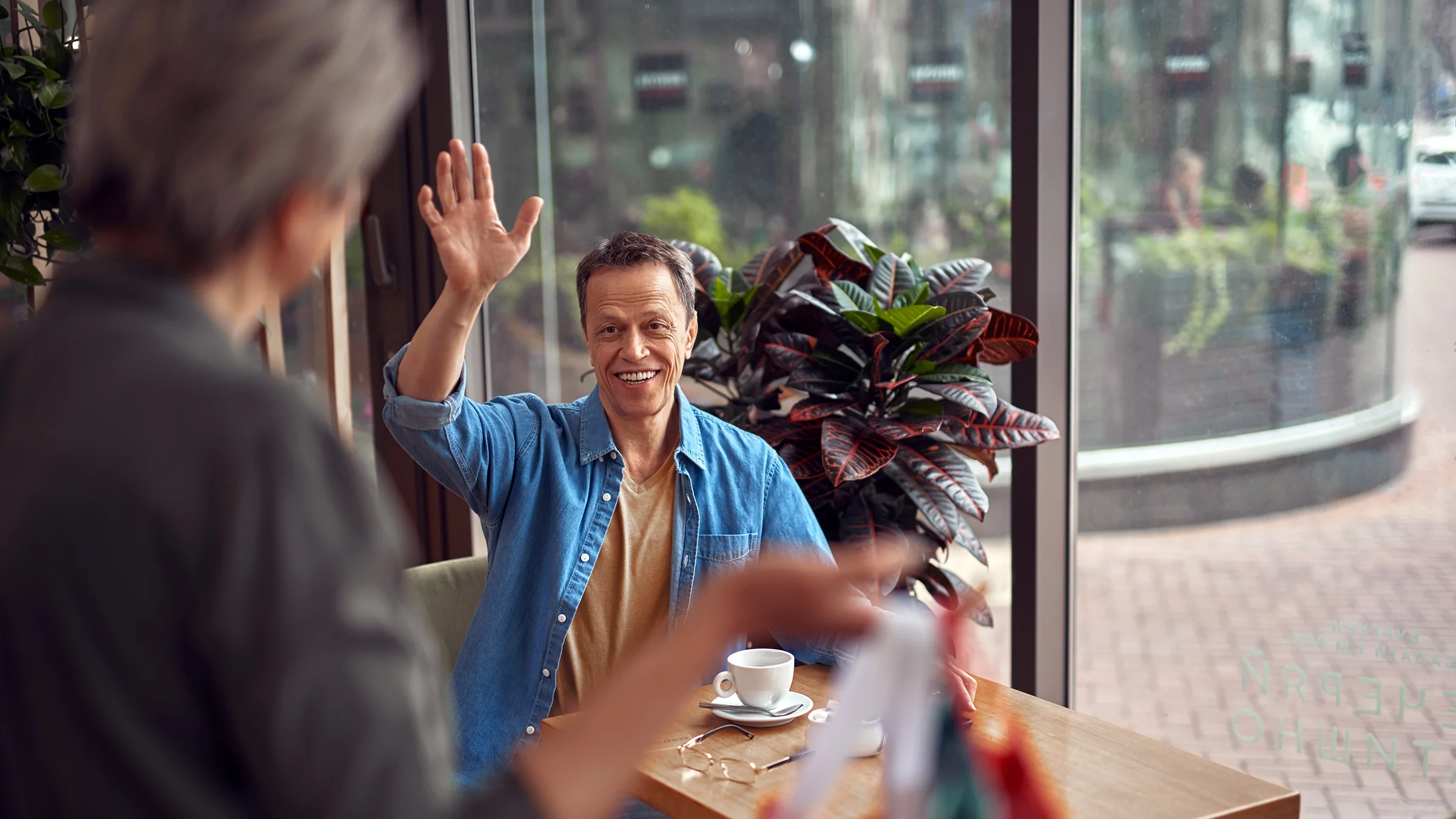 A man sits in a cafe and waves to a woman walking by.