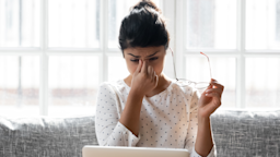 Woman taking off her glasses to rub her dry eyes. She is sitting at her laptop on the couch.
fizkes/iStock via Getty Images
