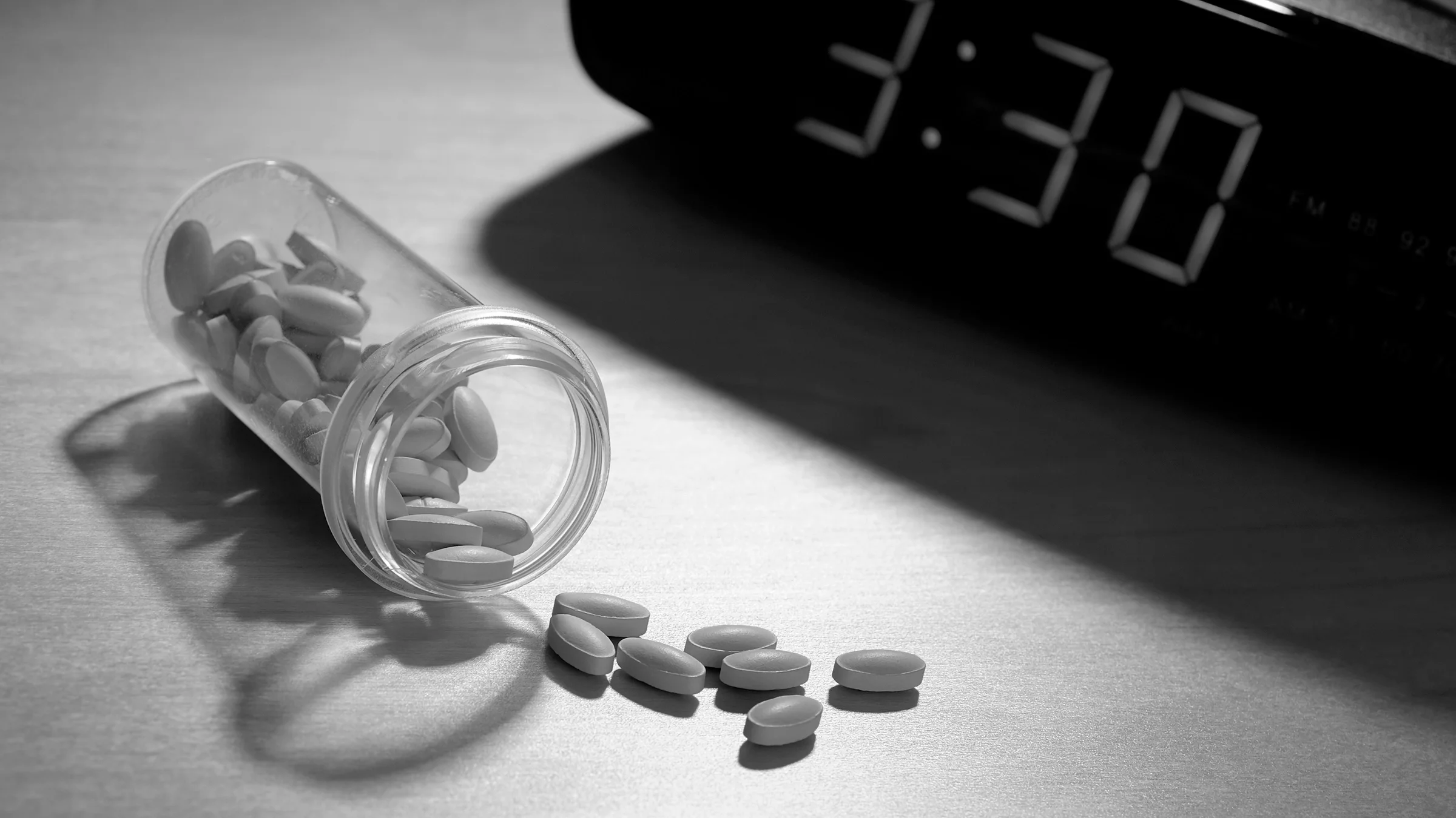 Black-and-white image of pills spilling out of a clear pill bottle next to an alarm clock reading 3:30.