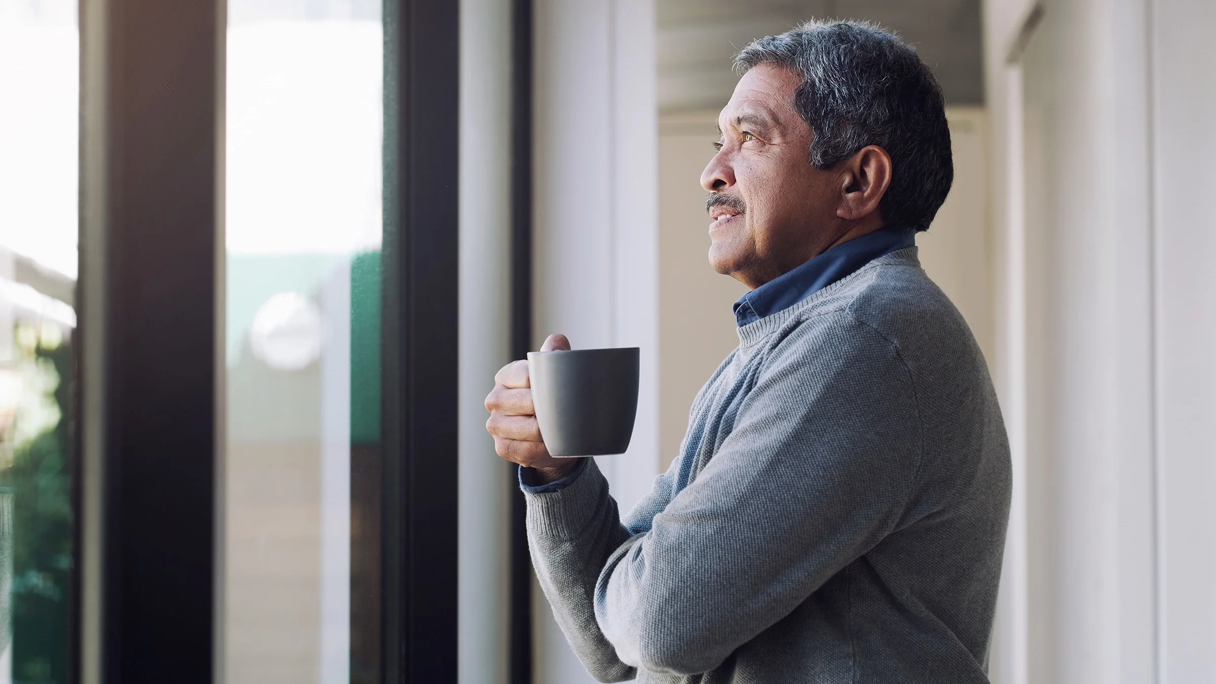 Older man holding a cup of coffee and looking out the window. He has a half smile on his face.