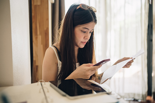 A woman using her phone while looking at paperwork in her house.