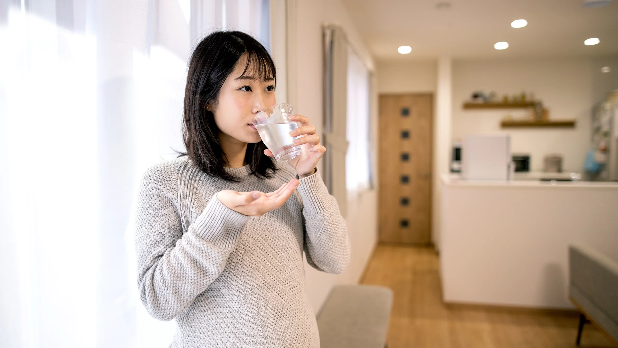 Pregnant woman taking medication with a glass of water. She is wearing a long-sleeve textured sweater.