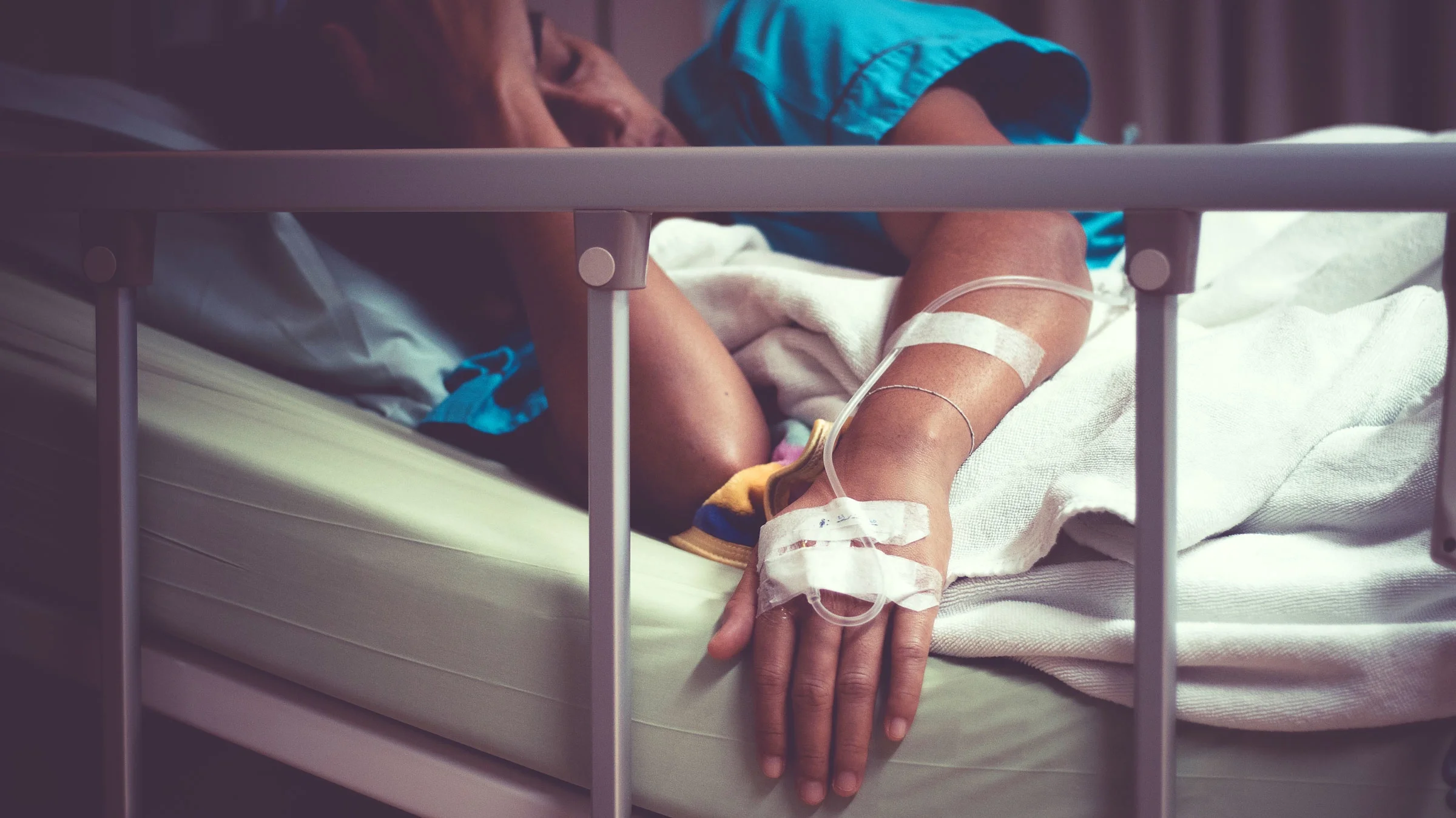 A person is sick and lying in a hospital bed with an IV in their hand.