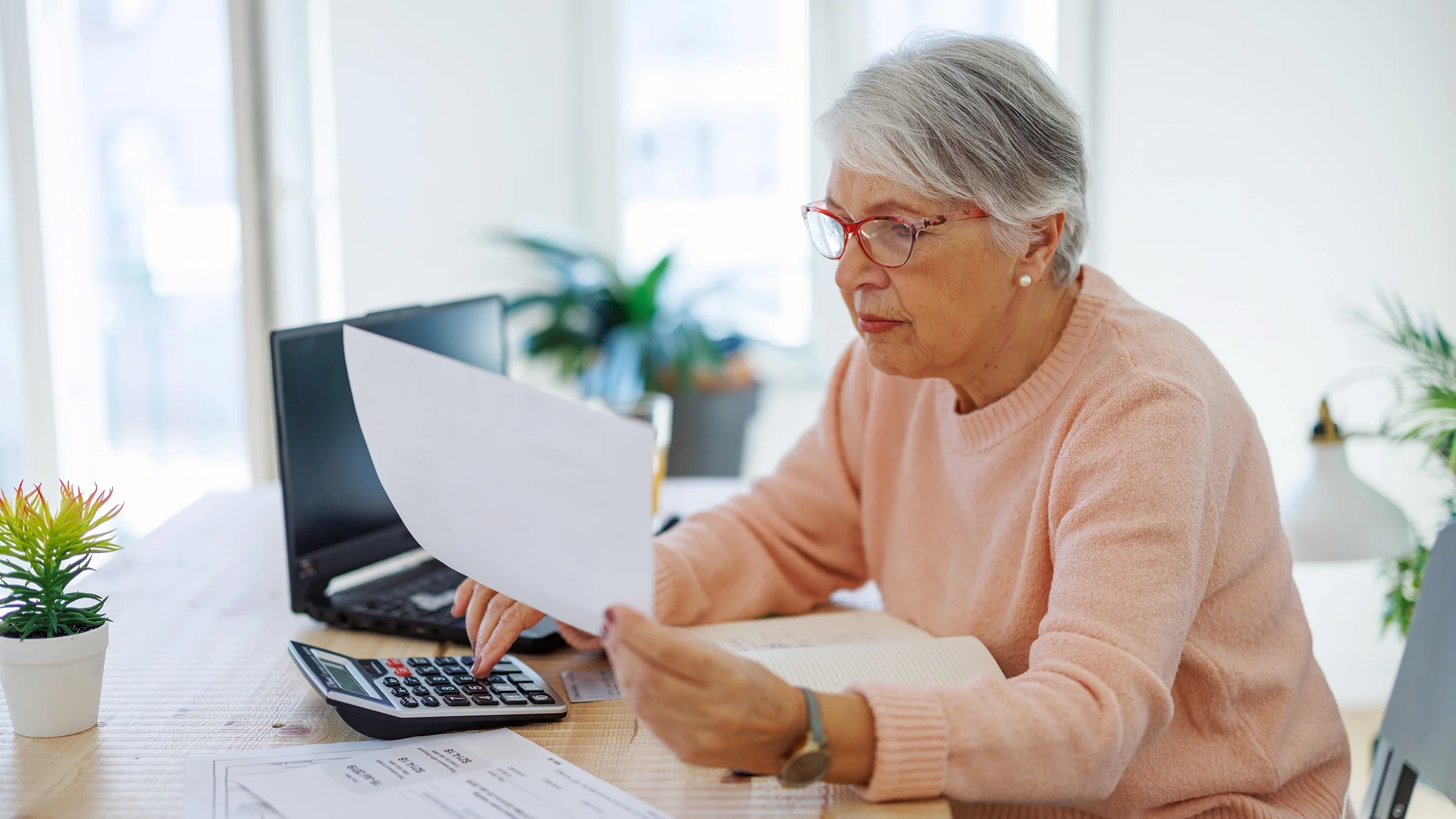 A senior woman sits at a table, using a calculator and looking at papers.