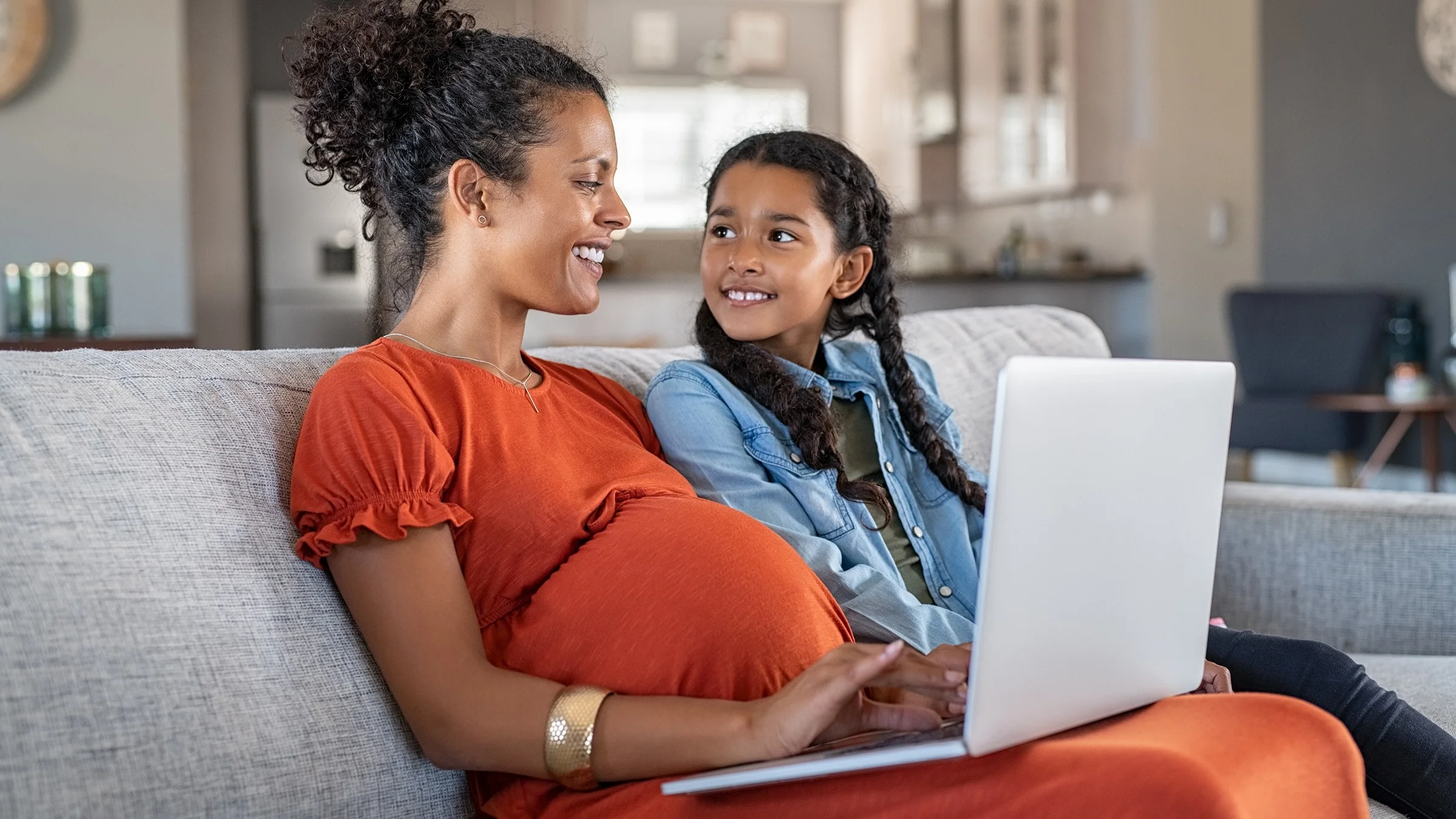 Young daughter and mother sitting on the couch. The mother is pregnant and wearing a red orange colored dress. She has a laptop on her lap and is typing while she smiles at her daughter.