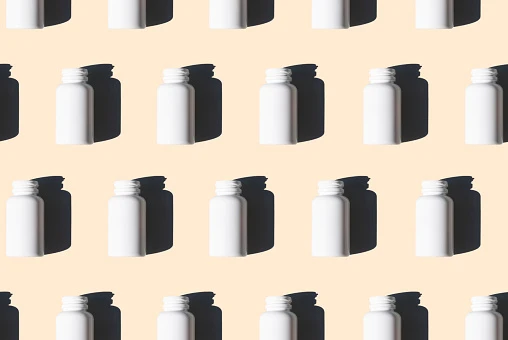 A pattern of white pill bottles on a tan or cream background with shadows falling on the background.