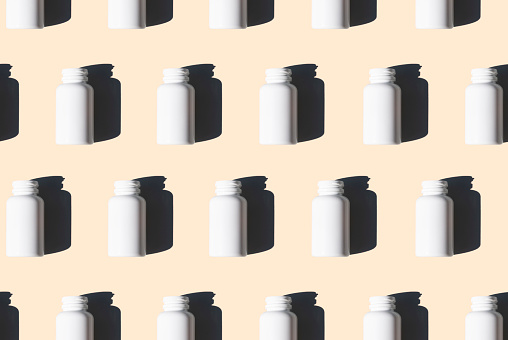 A pattern of white pill bottles on a tan or cream background with shadows falling on the background.