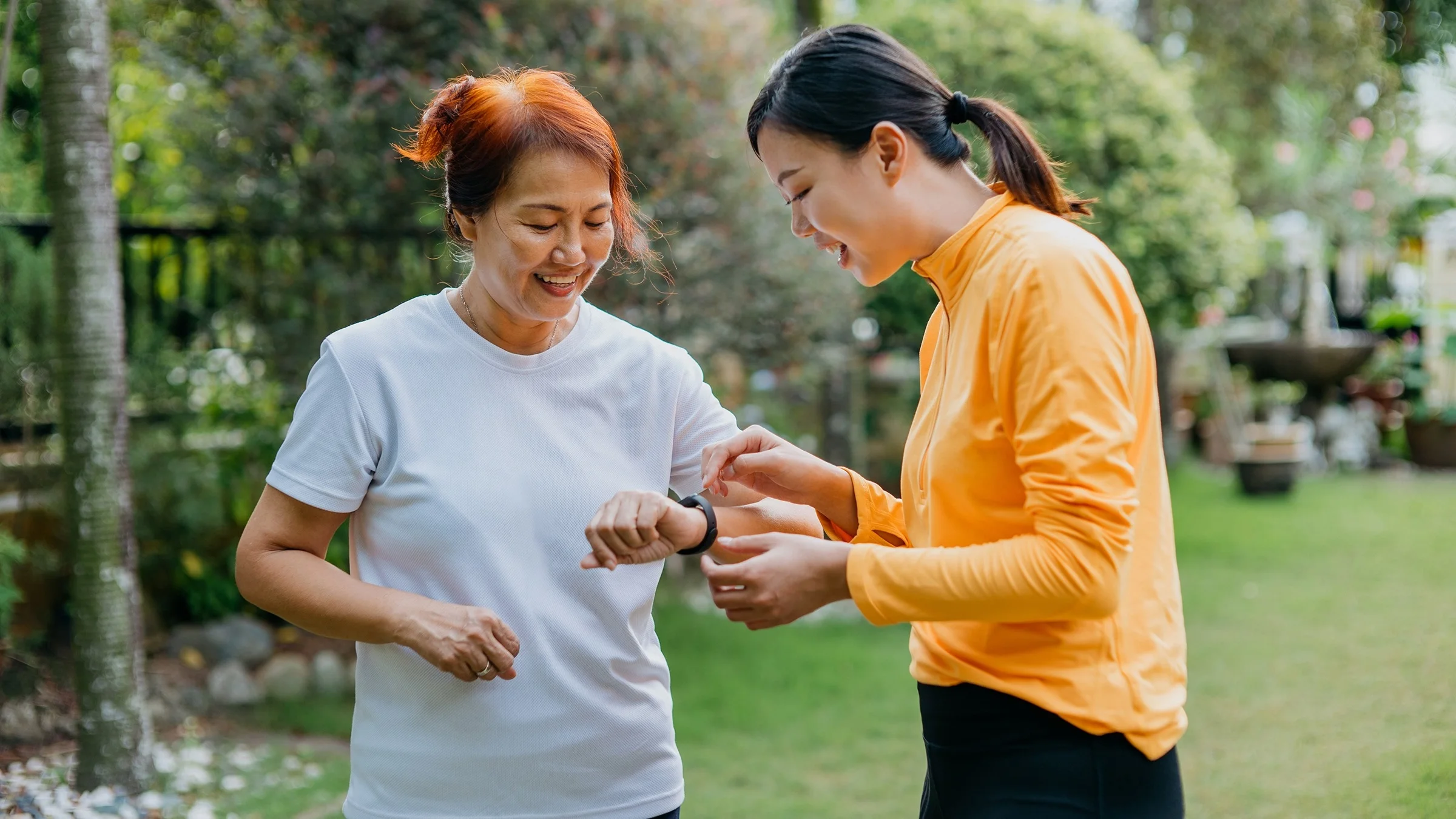 Adult daughter helping her older mother use a smart watch to track her heart rate. Both of them are in workout clothes outside.