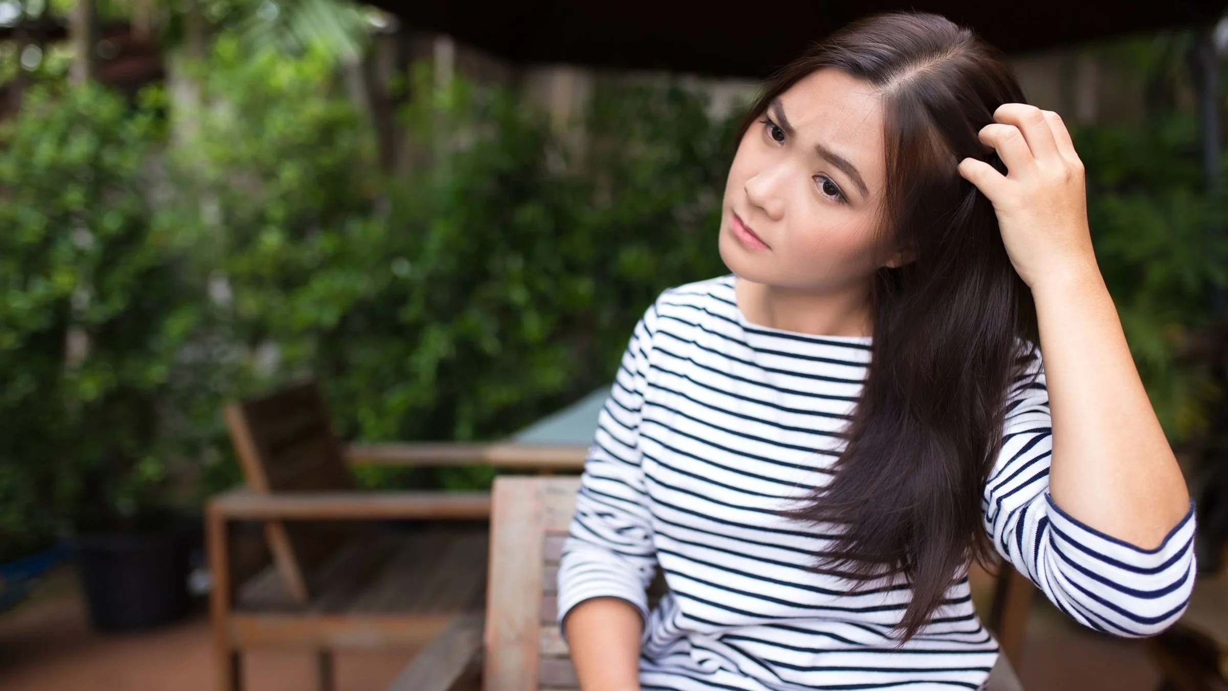 Portrait of a woman with long, dark hair scratching her scalp. She is outside on a patio with a lush green background.