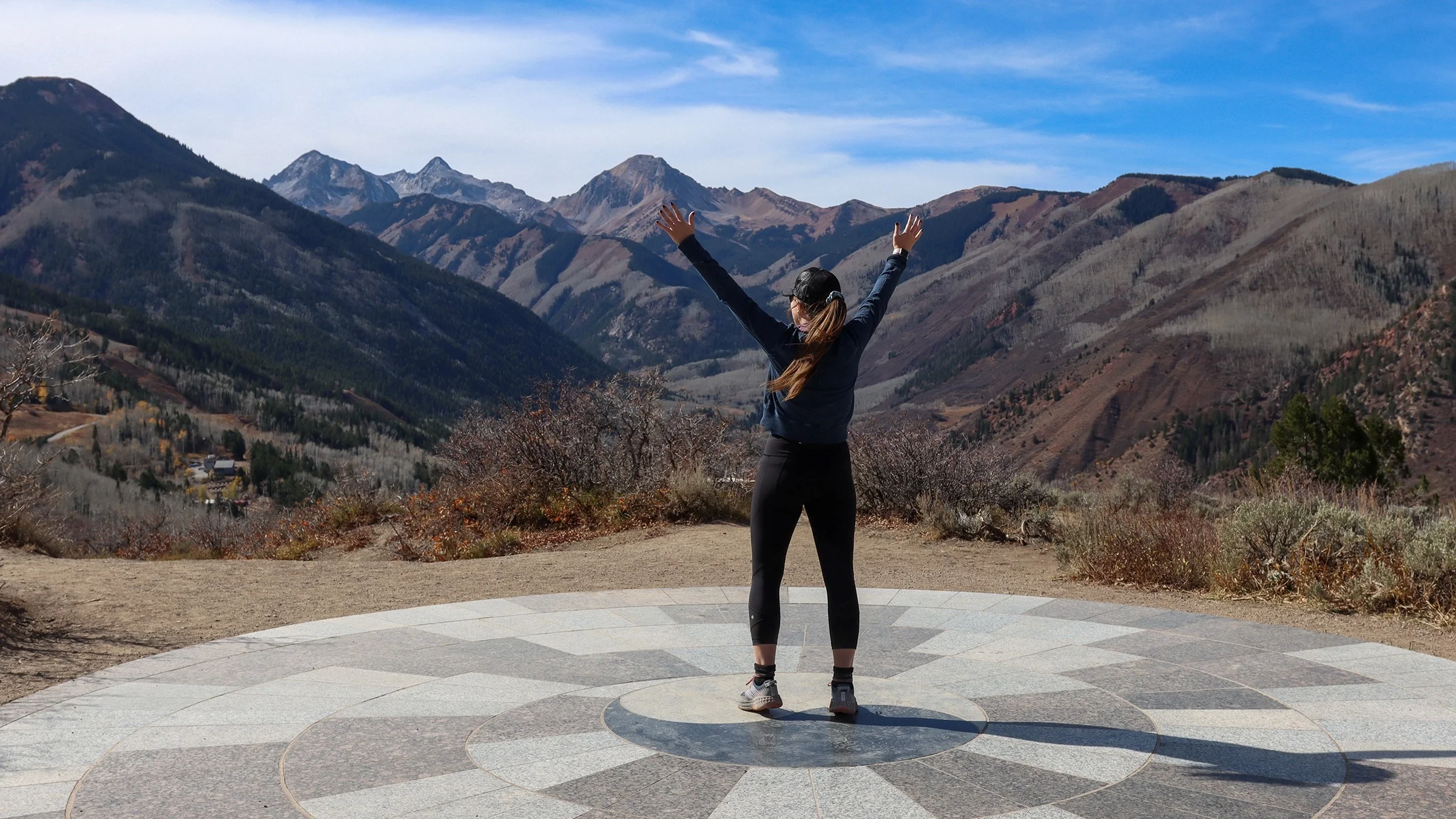 Woman standing outside with arms raised facing mountains.