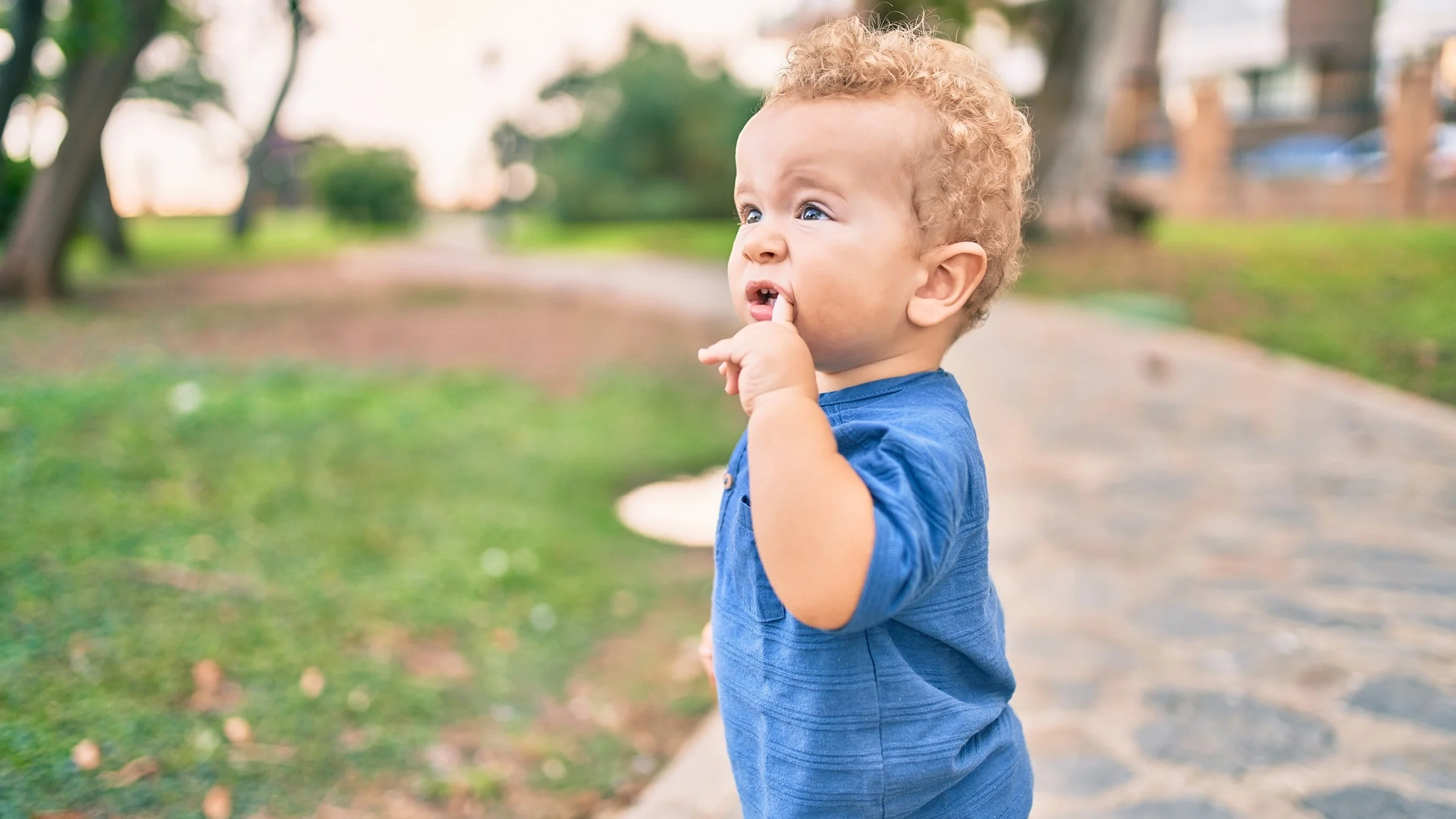 Little toddler with his finger in his mouth while at the park. He has cute curly hair and a a pained look on his face from a tooth ache. He is looking up with his eyebrows raised at someone out of frame.