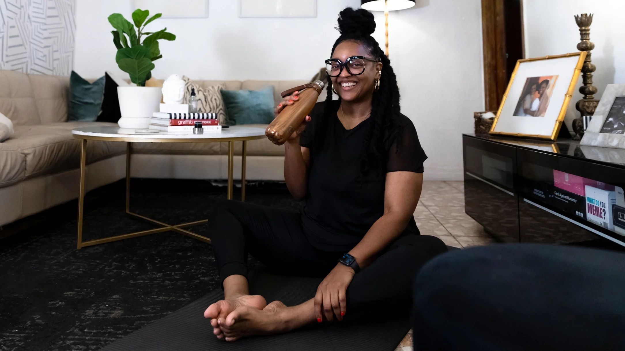 Nikki Nurse sitting down to practice yoga in her living room. She is holding a gold metal water bottle.