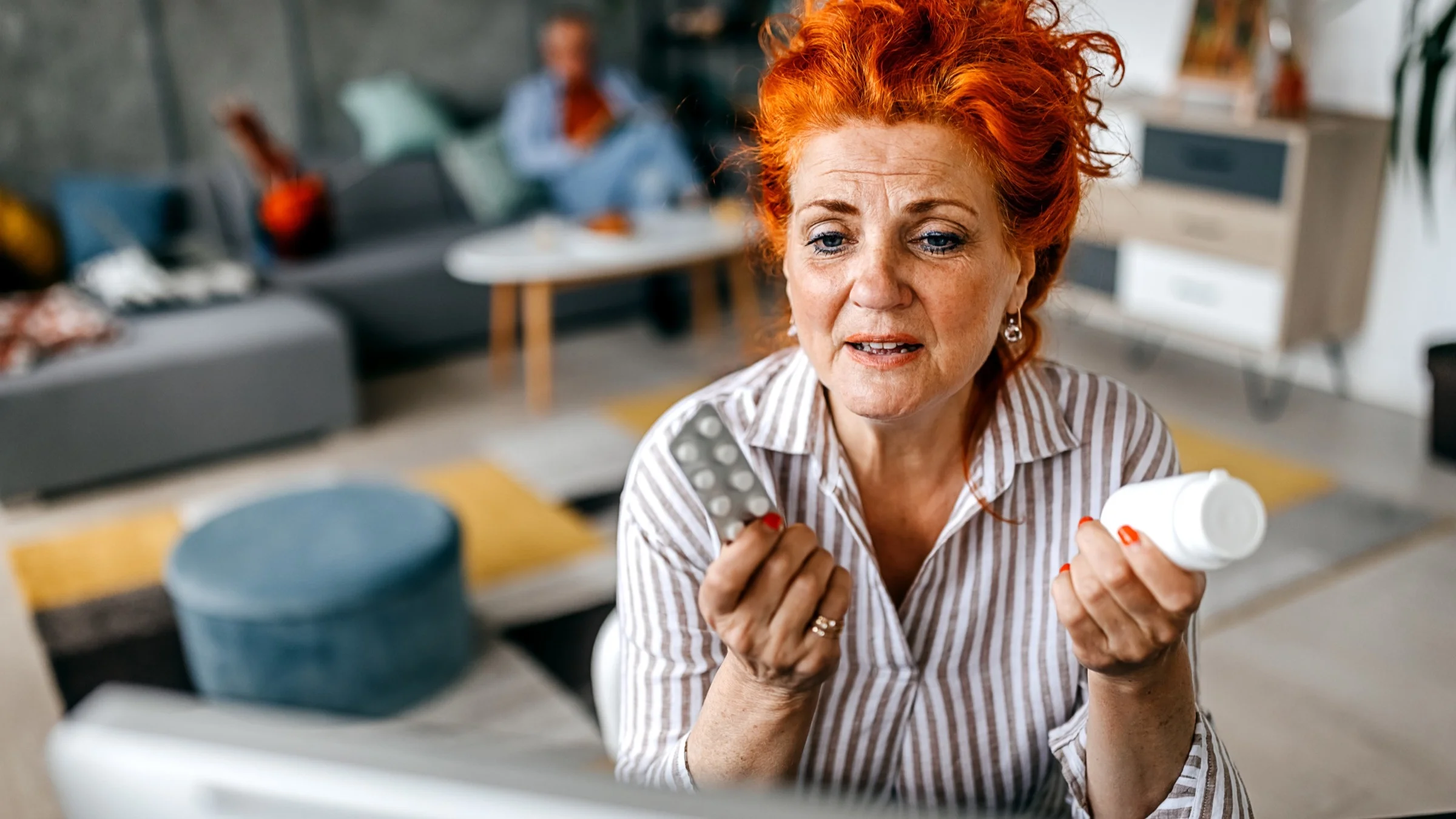 Close-up of a senior woman, with bright orange hair, showing her doctor her medications on a telehealth visit at her computer.