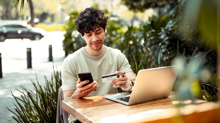 A man with his laptop at an outdoor cafe, making an online card payment on his phone.