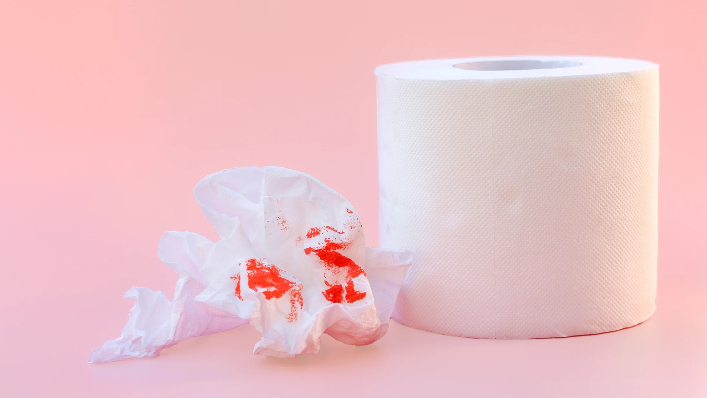 There’s a toilet paper roll with a crumpled bit next to it with red dye implying blood. The background is light pink.